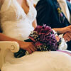 Violett lila Brautstrauß auf dem Schoß der Braut, händchenhaltend mit dem Bräutigam.